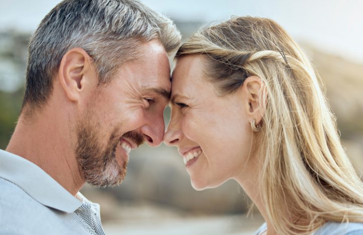 Happy and loving mature caucasian couple enjoying a romantic date at the beach together on a sunny day. Cheerful affectionate husband and wife standing face to face with noses touching for eskimo kiss while bonding on vacation outdoors